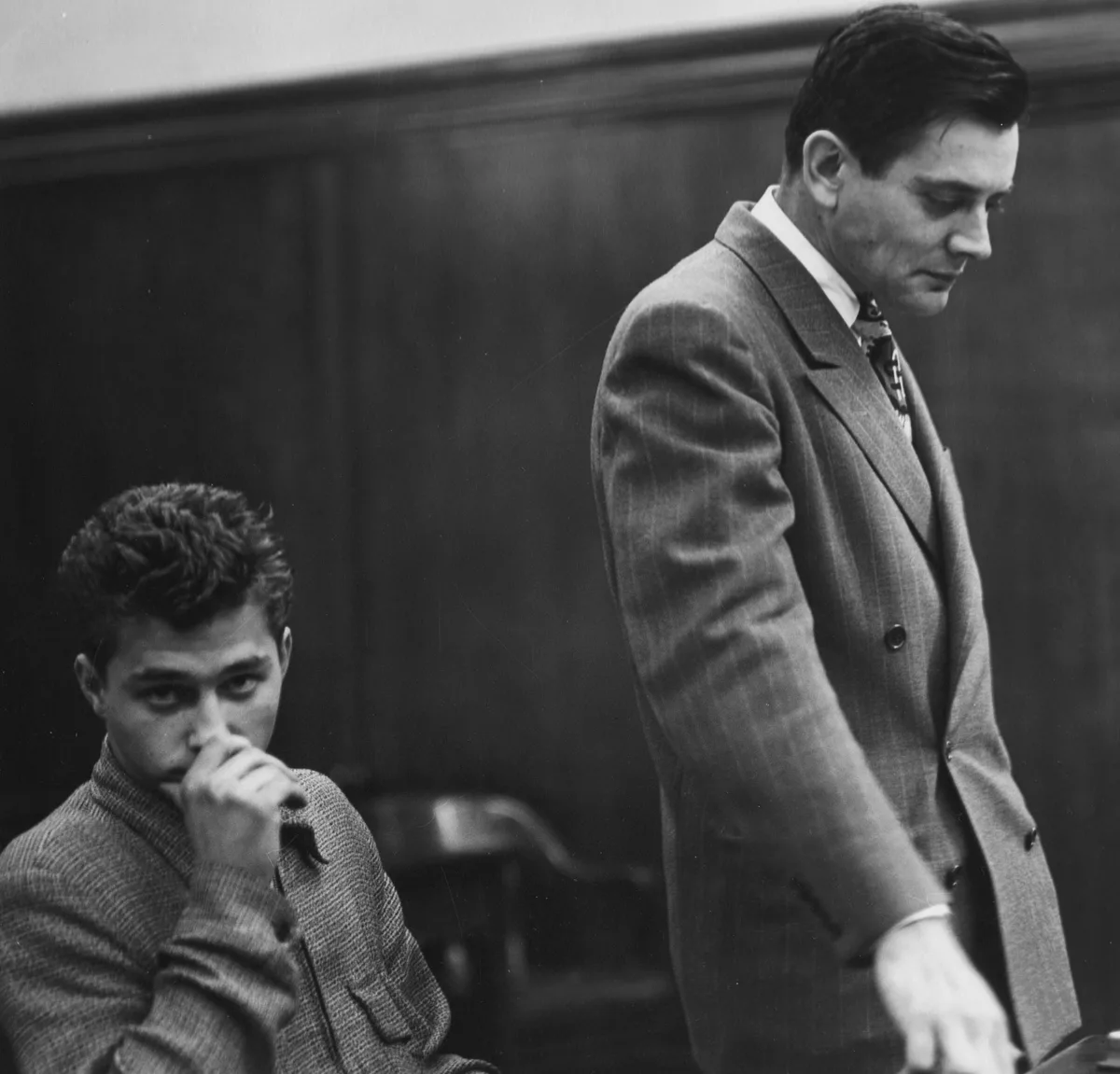A young defendant sits with his hand over his mouth while his lawyer stands beside him, looking down at the table