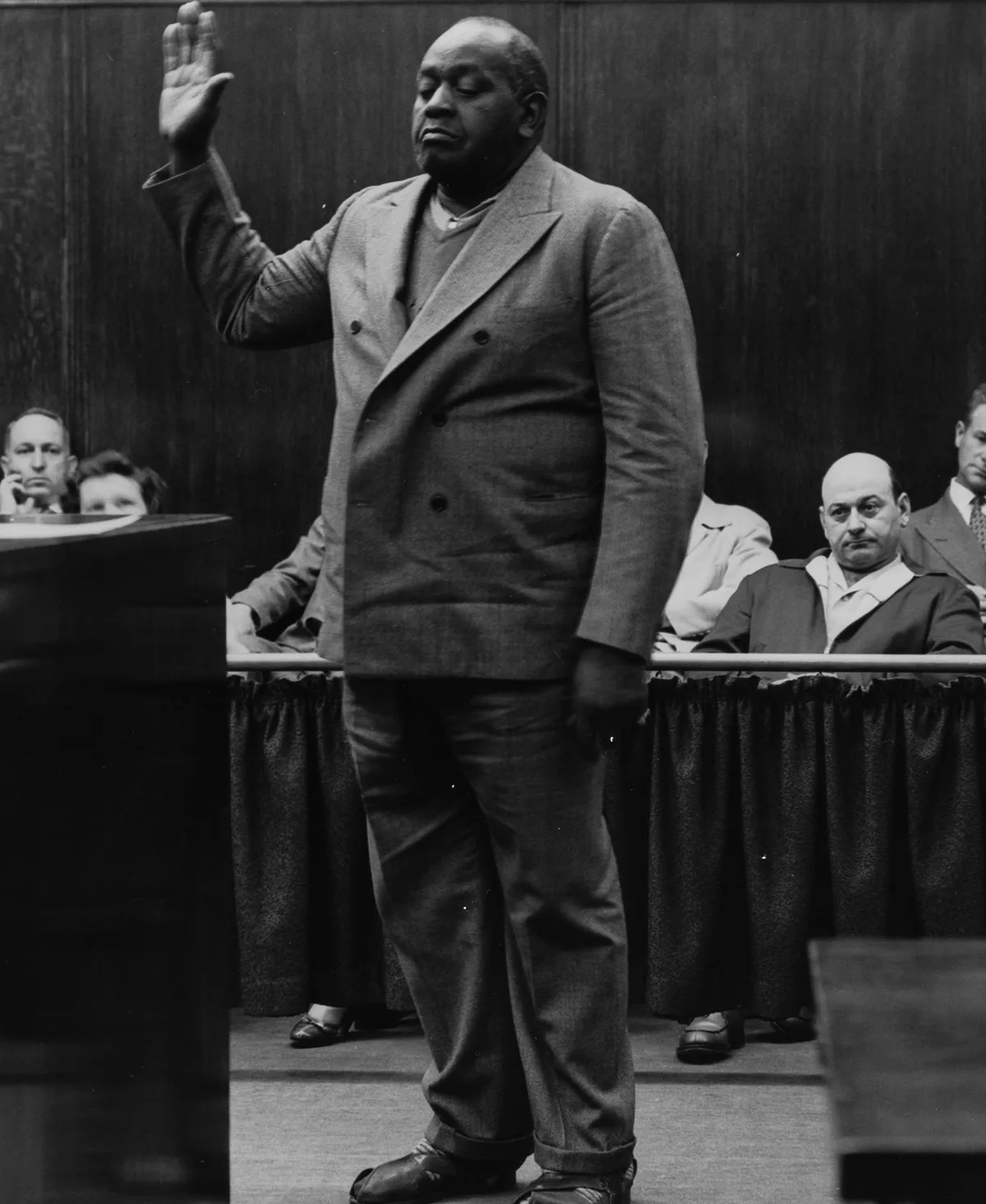A man in a double-breasted suit raises his right hand to take the oath, onlookers watching from behind a railing