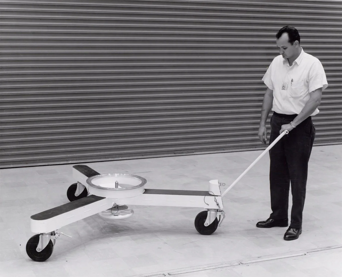 A technician in shirtsleeves and tie wheels a strange three-armed instrument cart across an empty floor