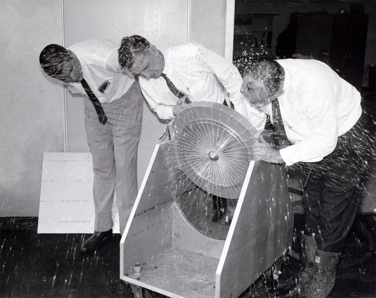 Three men in shirts and ties bend close over a spinning disc apparatus as it sprays water over them